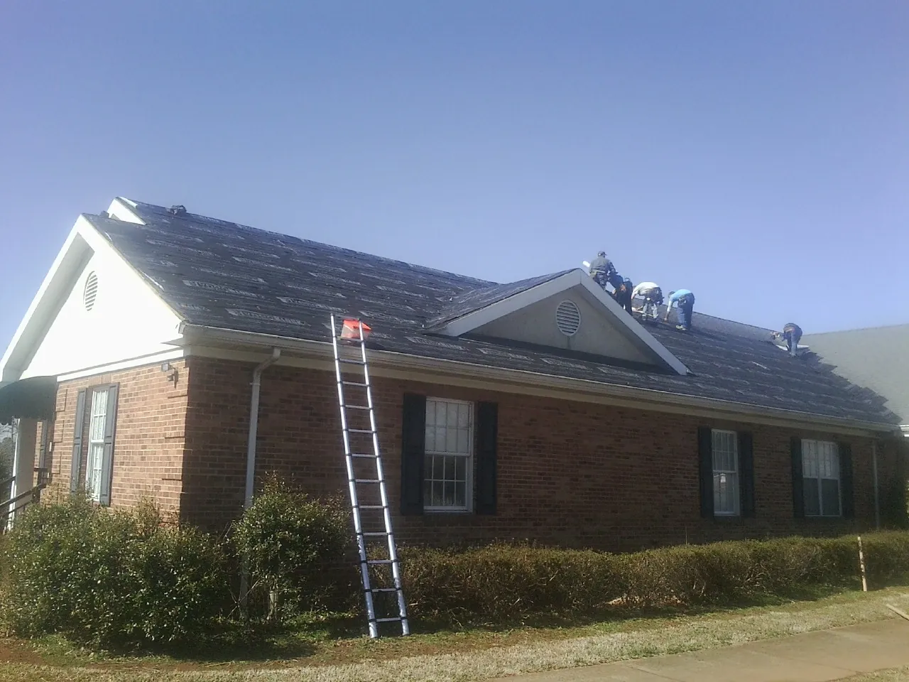 Crew tearing shingles off a brick ranch roof with a ladder set — Metal Roof Repair work by our crew in Selkirk