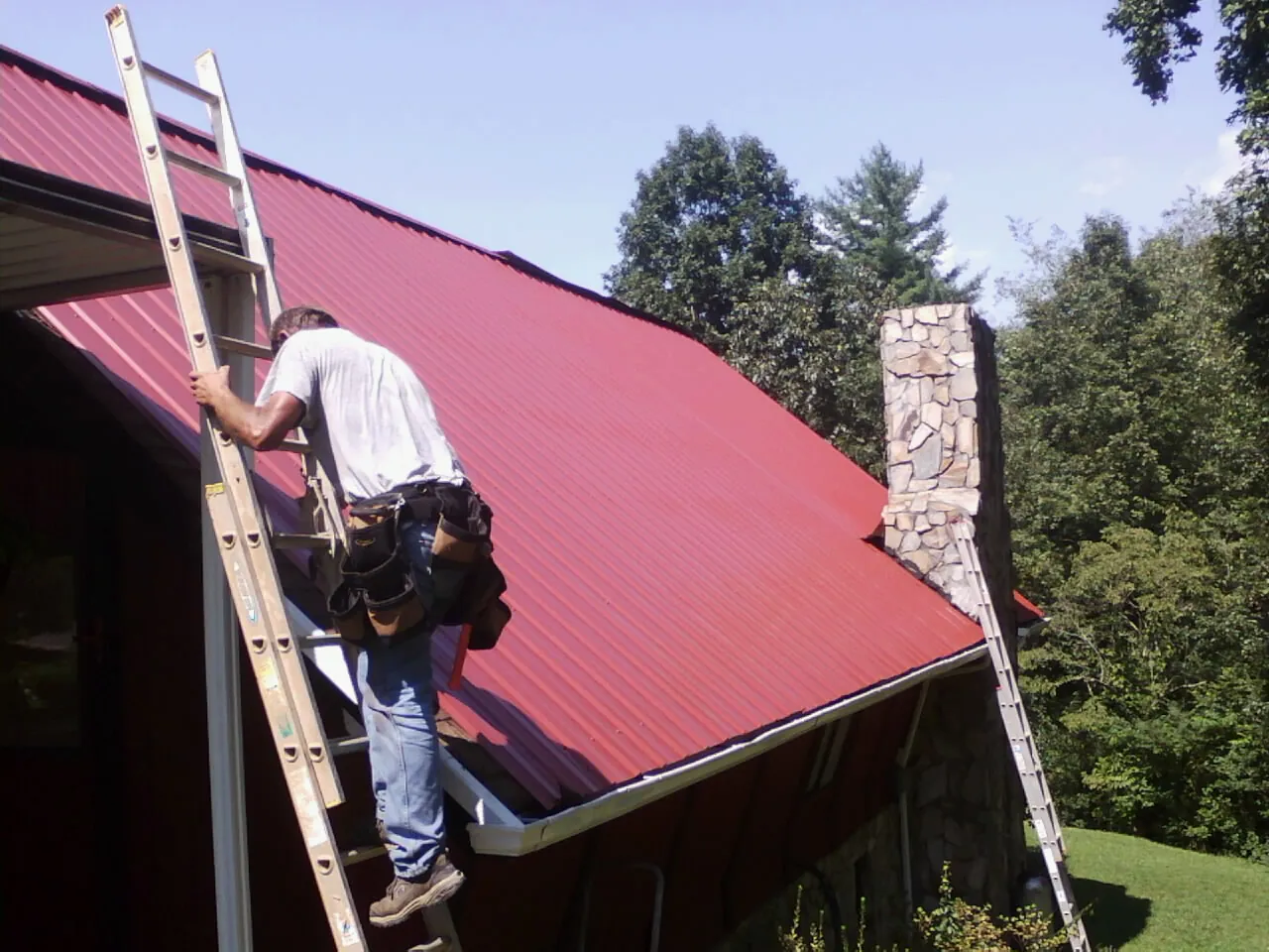 Roofer on a ladder beside a red metal roof and stone chimney — Soffit & Fascia Repair by Roofing Brothers in Selkirk